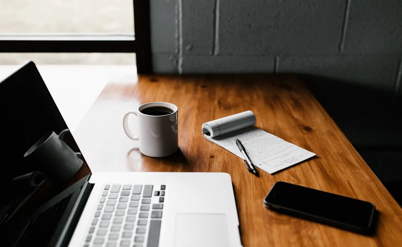 Escritorio con laptop, café y cuaderno junto a una ventana